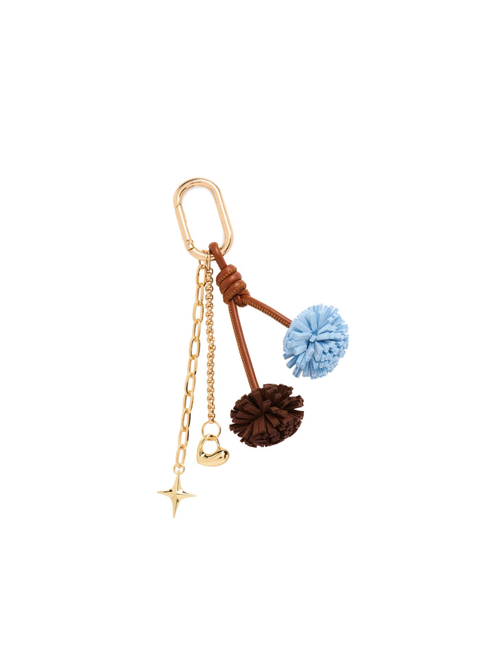 A gold-tone keychain or bag charm featuring a brown leather cord with two knotted pom-poms (one light blue, one dark brown), a gold-tone chain, a small gold bell, and a star-shaped charm, all attached to a gold carabiner.