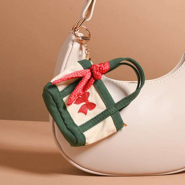 A small canvas keychain bag charm shaped like a gift box, featuring dark green fabric sides and handles, a cream canvas front, and a red fabric bow with white polka dots, hanging from a gold ring on a white handbag.