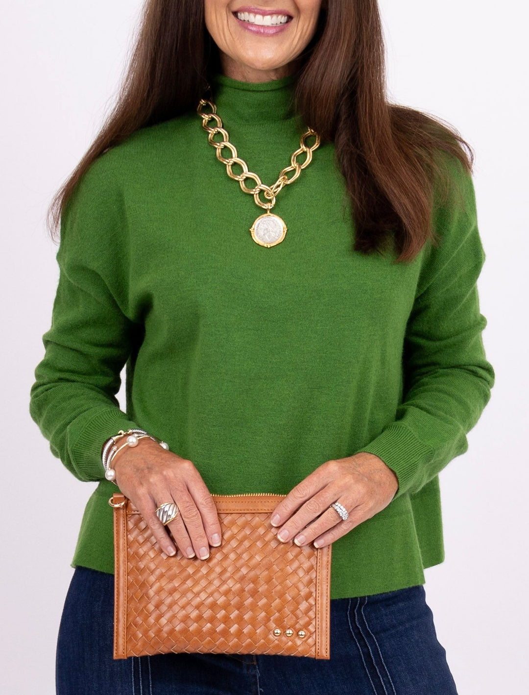 A woman wearing a green turtleneck sweater and holding a tan woven leather clutch, accessorized with a chunky gold chain necklace and rings, against a plain white background.