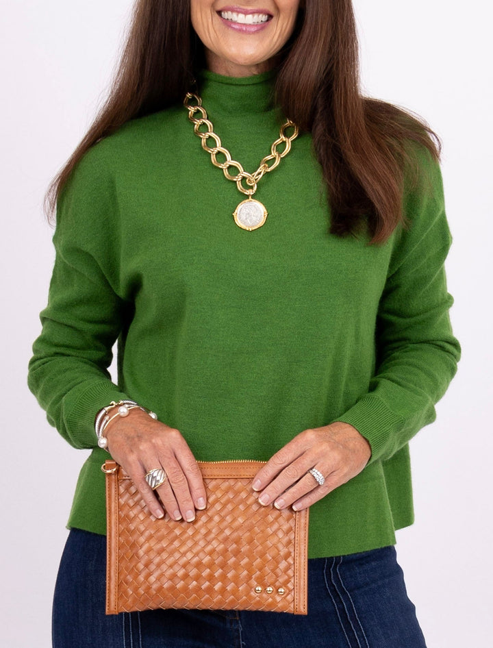 A woman wearing a green turtleneck sweater and holding a tan woven leather clutch, accessorized with a chunky gold chain necklace and rings, against a plain white background.