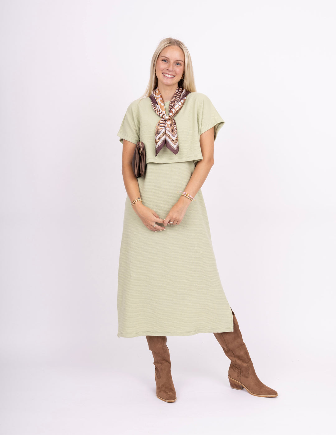 A woman modeling a light sage green two-piece set consisting of a short-sleeve top and matching midi skirt, styled with a brown and white patterned scarf, a small brown handbag, and brown suede boots against a white studio background.