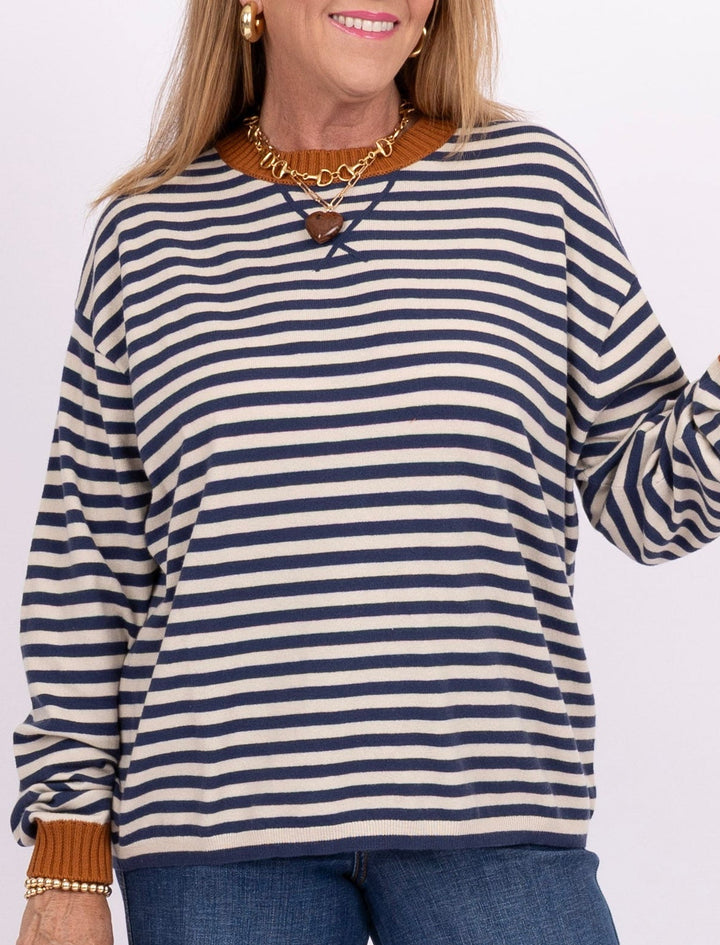 A woman modeling a navy and white horizontally striped long-sleeve top with brown cuffs, paired with wide-leg blue jeans, tan clogs, and holding a brown clutch against a plain white studio background.