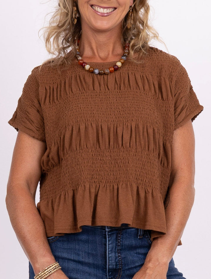A smiling blonde woman stands against a plain white studio background wearing a brown ruffled short-sleeve top, blue jeans, and olive green sneakers, while holding a small cream clutch.