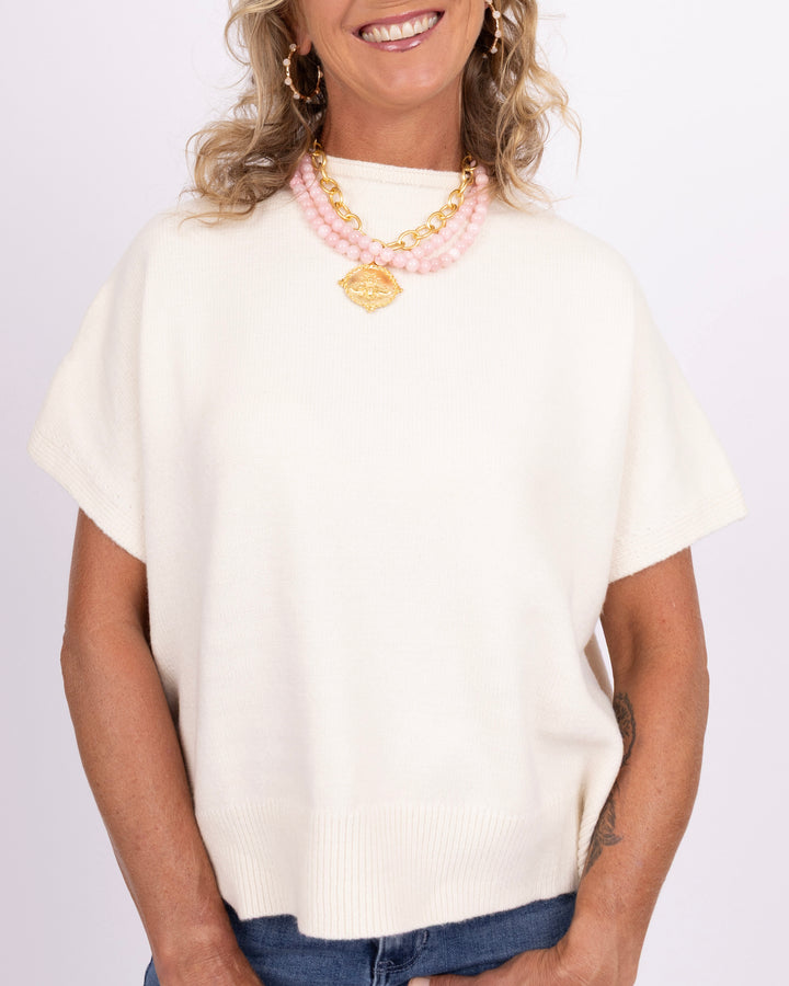 A woman wearing a cream-colored short-sleeve knit sweater with a ribbed hem, styled with a pink and gold beaded necklace and blue jeans against a light background.