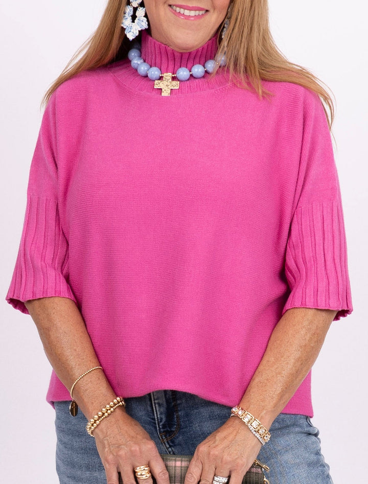 A woman modeling a bright pink short-sleeve top with blue jeans, holding a plaid clutch bag against a plain white studio background.