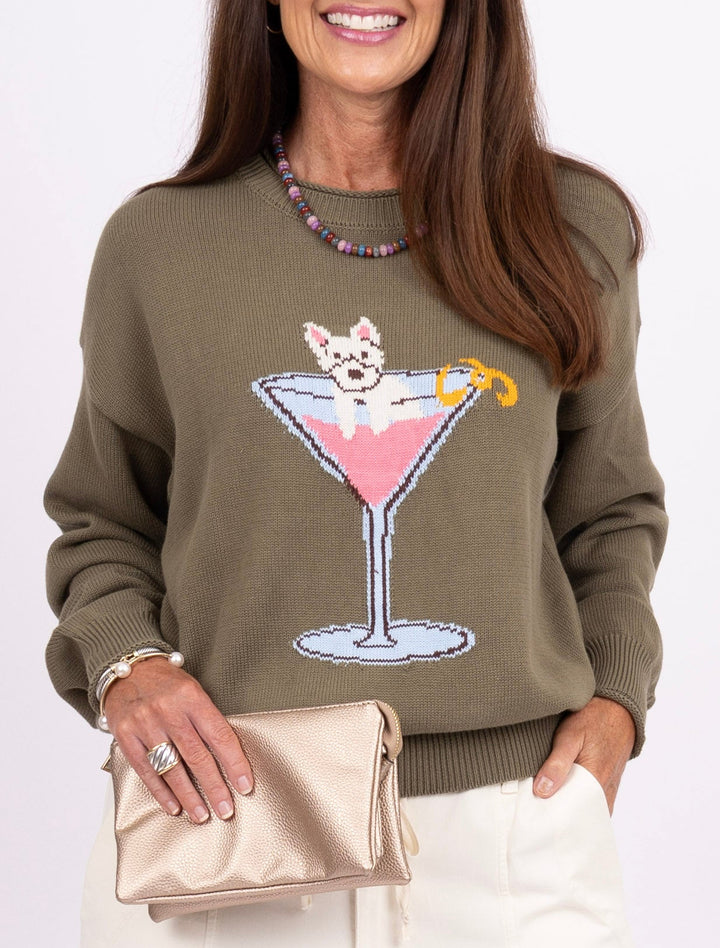 A woman modeling an olive green crewneck sweatshirt with a cocktail graphic, paired with white wide-leg pants, a metallic gold clutch, and pink sneakers, set against a plain light gray studio background.