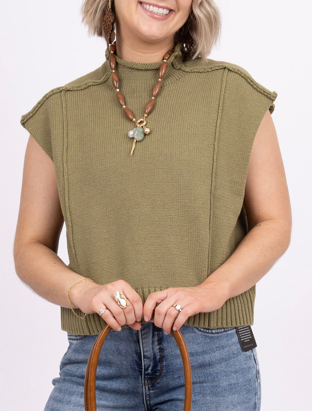 A woman modeling a sleeveless olive green top, blue jeans, and holding a brown leather handbag against a plain white studio background.