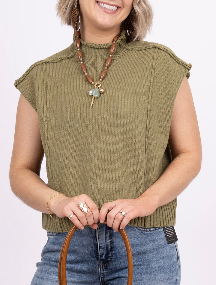 A woman modeling a sleeveless olive green top, blue jeans, and holding a brown leather handbag against a plain white studio background.