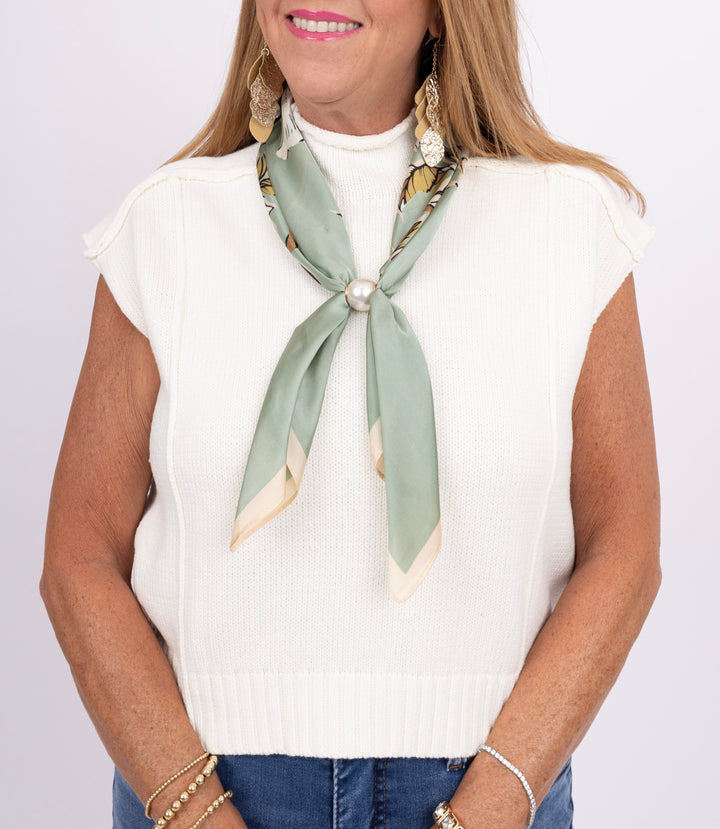 A woman wearing a white ribbed knit sleeveless sweater vest, styled with a sage green and beige patterned silk neck scarf tied in a bow at the collar, accessorized with gold earrings and bracelets.
