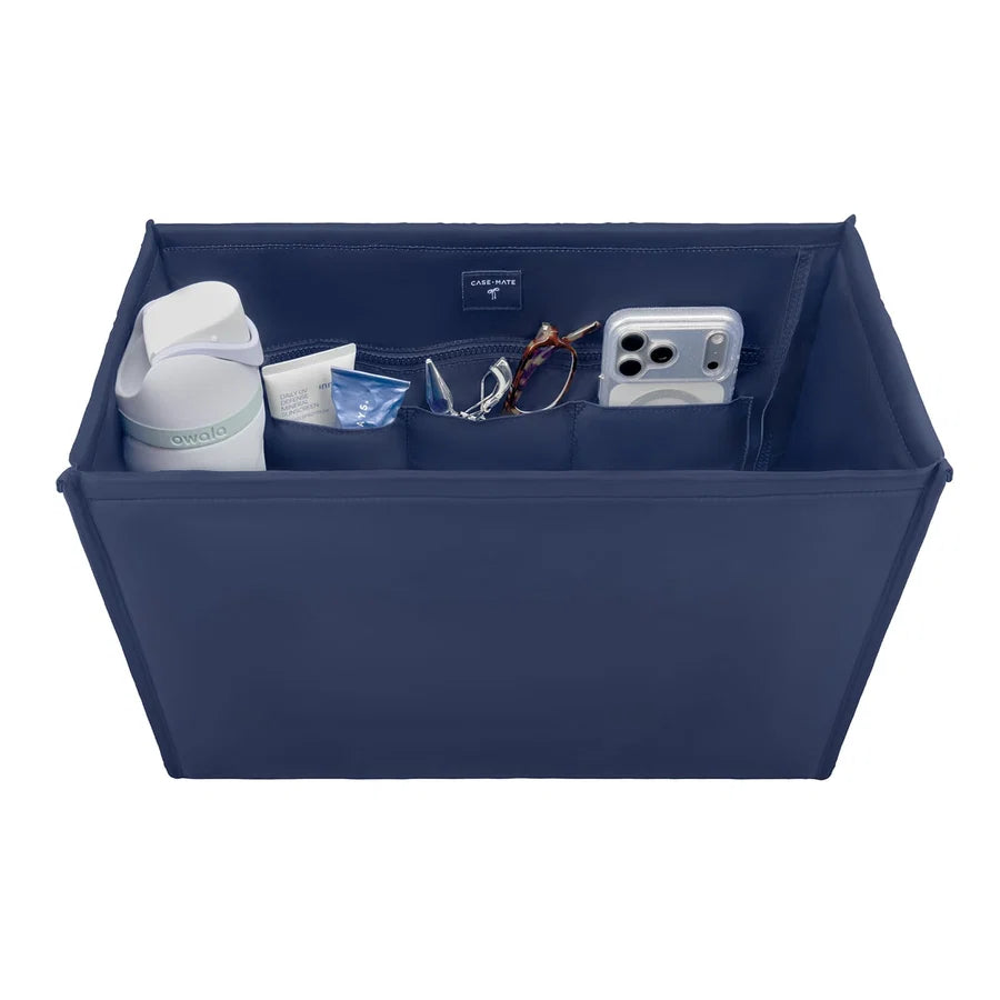 A navy blue fabric organizer tray with multiple interior pockets holding a white bottle, a small blue item, eyeglasses, and a silver smartphone, shown on a plain white background.