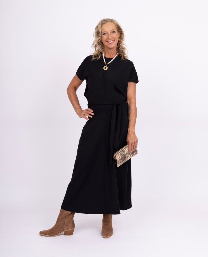A woman modeling a black short-sleeve midi dress with a tie waist, paired with brown ankle boots and a plaid clutch, against a plain white studio background.