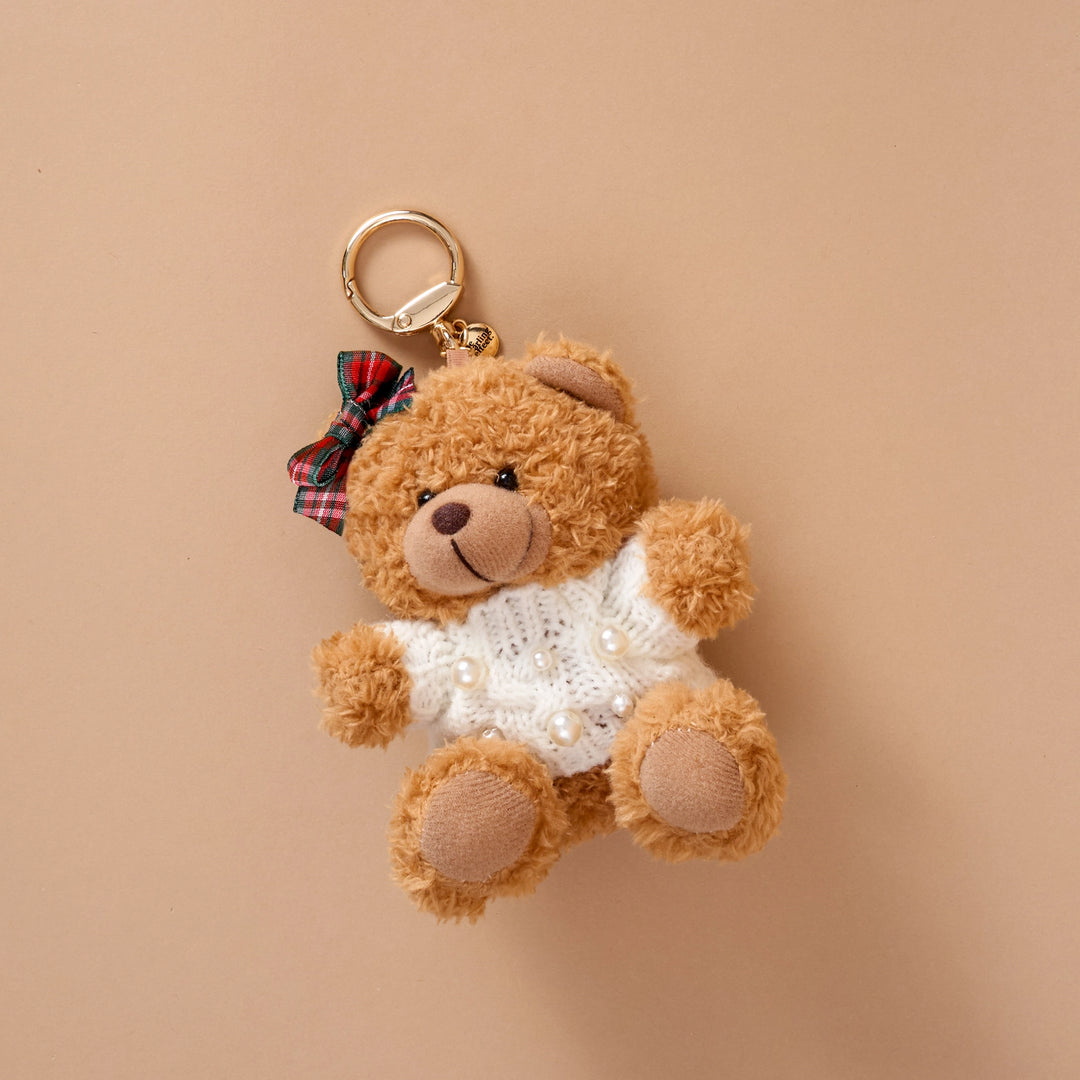 A small plush teddy bear keychain in warm brown, wearing a white knit sweater with pearl details and a red plaid bow, attached to a gold key ring against a solid beige background.