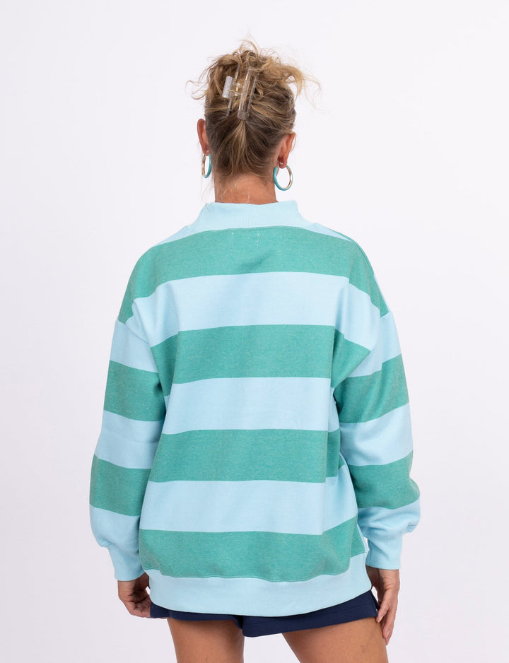 A woman is shown from behind wearing a light blue and mint green horizontally striped long-sleeve sweatshirt, a dark navy mini skirt, and pink sneakers against a plain white studio background.