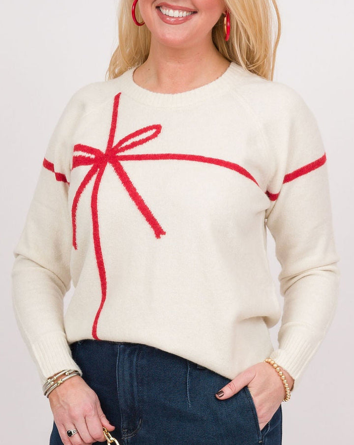 A woman modeling a cream knit sweater with a red bow graphic on the chest, paired with dark blue wide-leg jeans, a bright red clutch, and brown shoes, against a light gray studio background.