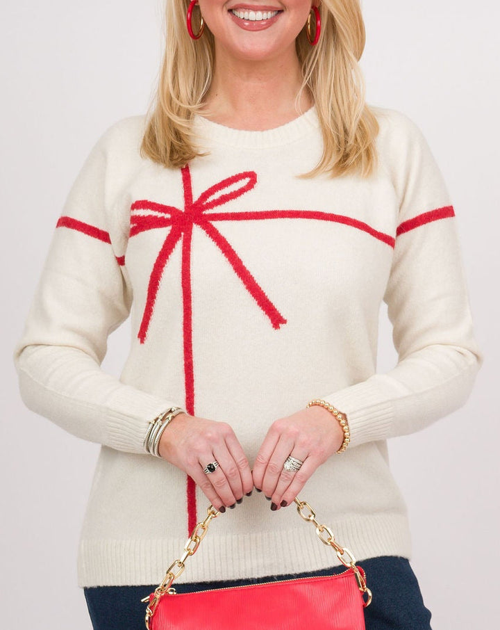 A woman modeling a cream knit sweater with red trim and a red bow motif, paired with dark blue wide-leg jeans and holding a small red handbag against a light gray studio background.