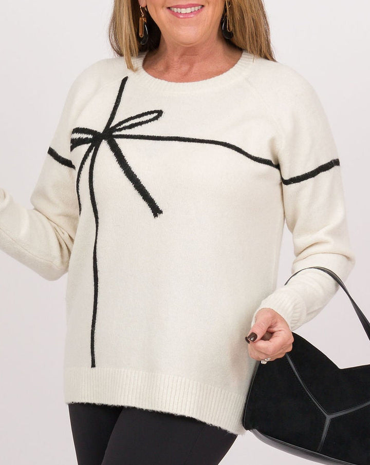 A woman modeling a cream knit sweater with black trim and a bow detail, paired with black leggings and black knee-high boots, holding a black handbag against a light gray studio background.