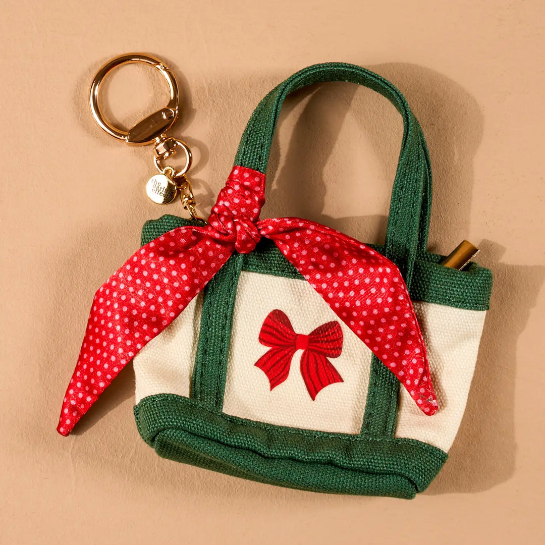 A small fabric tote keychain bag with green canvas trim, a white front panel, a red bow motif, and a red-and-white polka dot scarf tied around the handle. It has a gold-tone key ring and a pen slot on the side, photographed on a warm beige background.