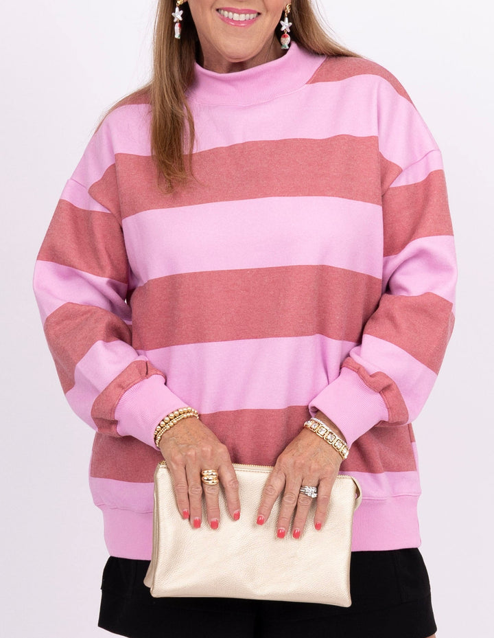 A woman modeling a pink and light pink striped mock-neck sweatshirt, paired with black shorts, a cream clutch, and clear wedge sandals, against a plain white studio background.