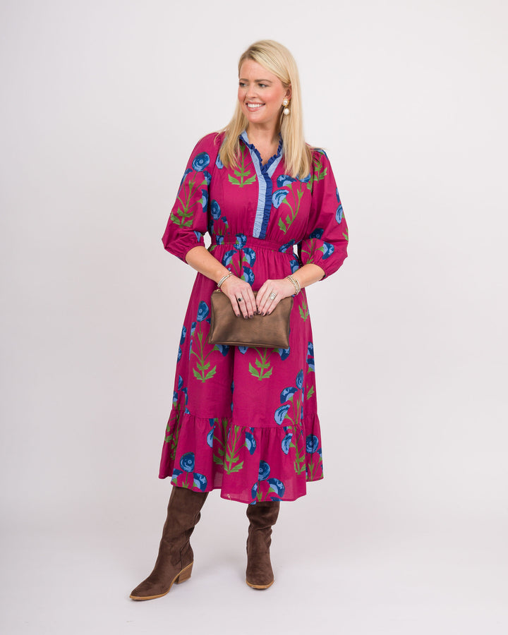 A woman modeling a bright pink tiered midi dress with a blue and green floral print, holding a brown clutch bag and wearing brown boots against a light gray studio background.