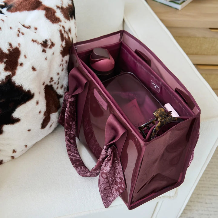 A translucent burgundy zippered organizer bag with patterned fabric handles, shown open on a white sofa next to a cow-print pillow.