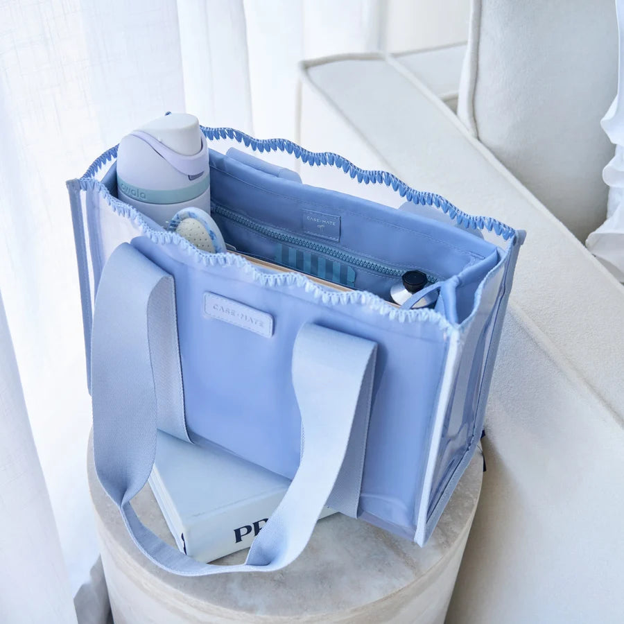 A light blue tote bag with white handles sits on a round stool in a bright, softly lit room. The bag is shown open, filled with travel items, and has a small white label on the front.