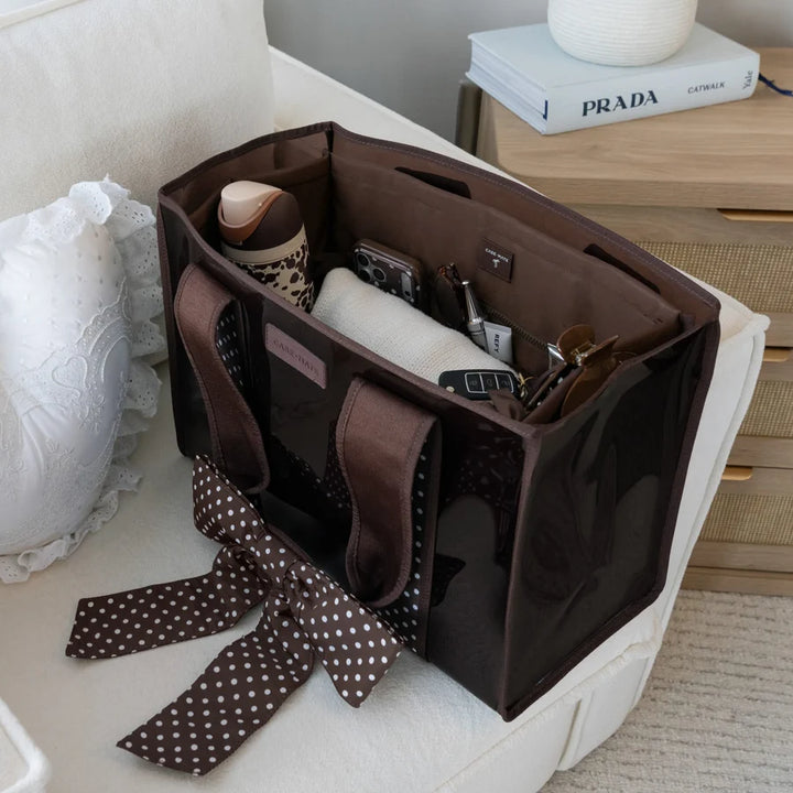 A brown tote organizer bag with a glossy finish and matching brown polka-dot ribbon handle, shown open on a white armchair with various accessories inside. A Prada book is visible on a wooden side table in the background.