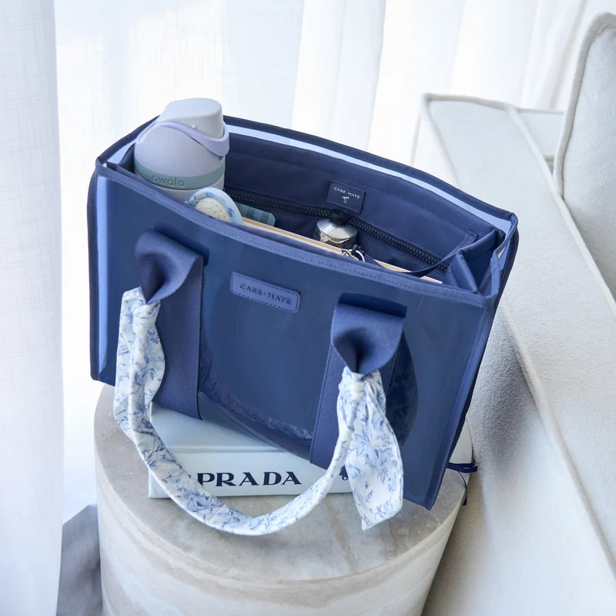 A navy blue diaper bag with patterned fabric handles sits on a round pedestal, with a bottle and baby items inside, in a softly lit indoor setting.