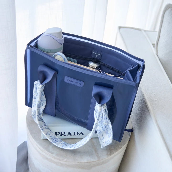 A navy blue diaper bag with patterned fabric handles sits on a round pedestal, with a bottle and baby items inside, in a softly lit indoor setting.