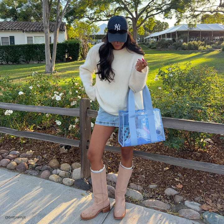 A woman stands outdoors on a sidewalk in front of a wooden fence and green lawn, wearing a cream sweater, denim shorts, beige knee-high boots, a black New York Yankees cap, and carrying a translucent light blue tote bag.
