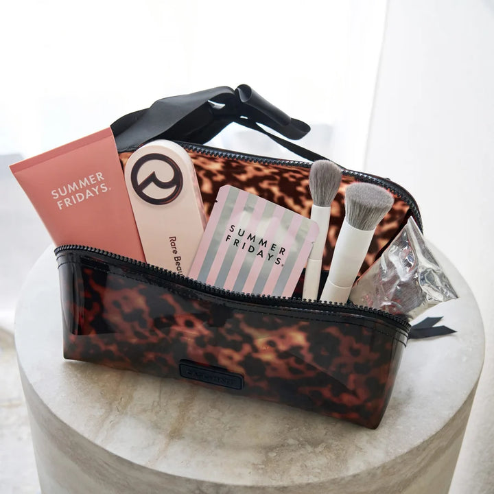A clear cosmetic pouch with a brown leopard print pattern and black trim, shown open on a round stone pedestal. Inside are makeup brushes, a pink tube labeled &