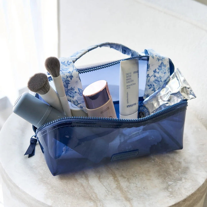 A translucent blue makeup bag with a blue and white floral pattern on the handles and interior lining, shown open on a round stone surface with makeup items inside.