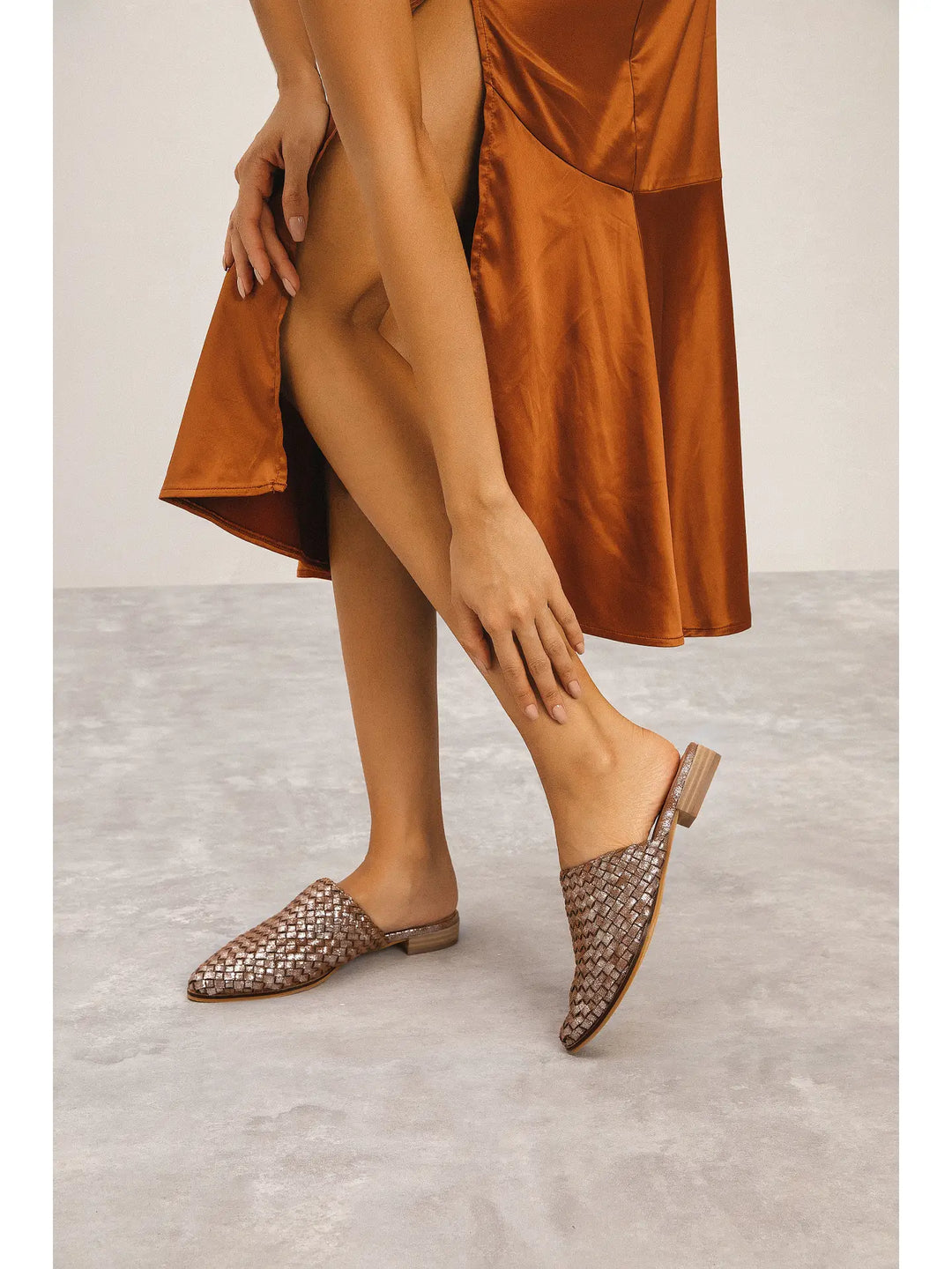A person wearing a brown satin dress and matching woven mule loafers, standing on a light gray floor against a neutral studio background.