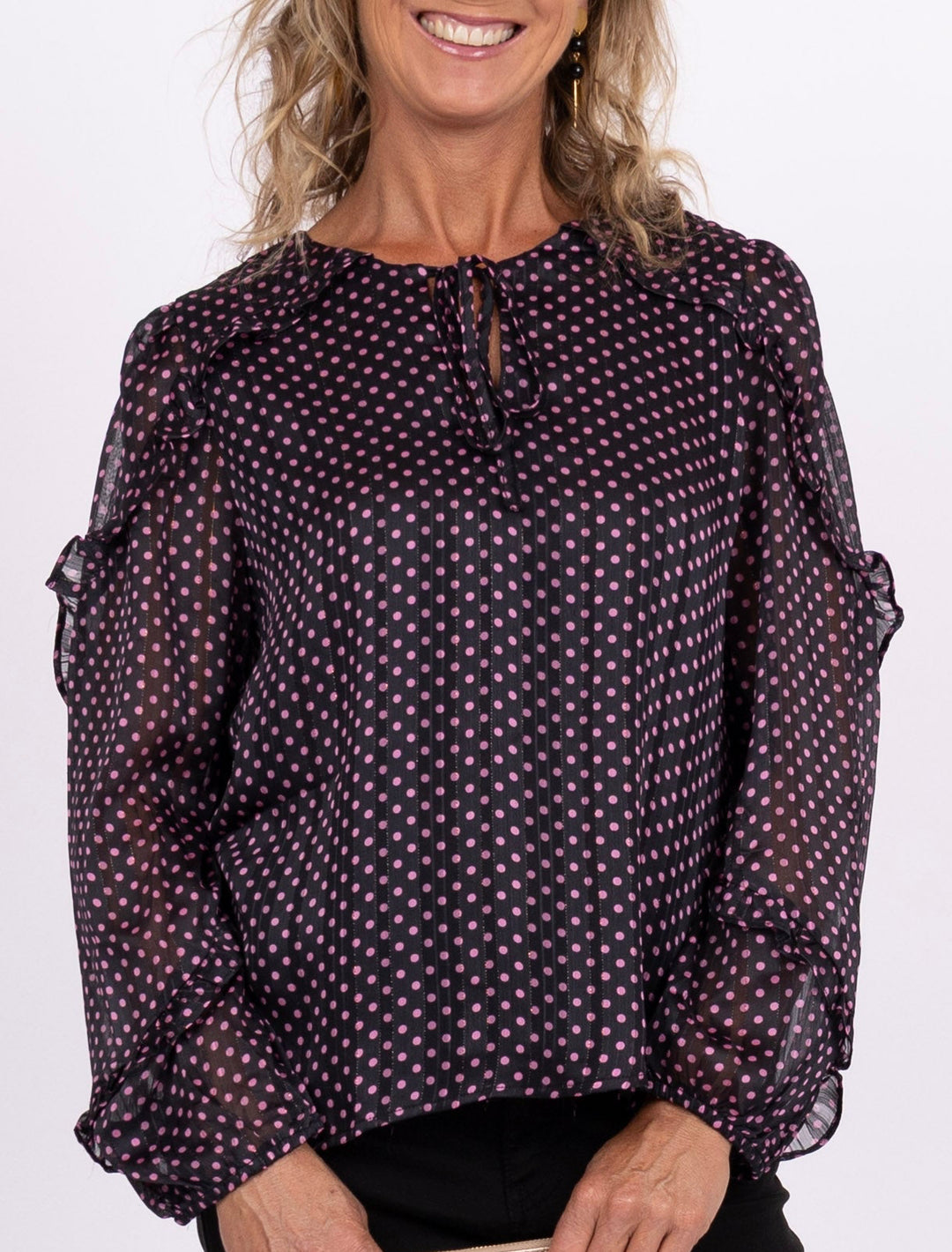 A smiling woman stands against a plain light background wearing a sheer long-sleeve blouse with a small repeating pattern, black pants, and pink sneakers, while holding a small cream clutch.