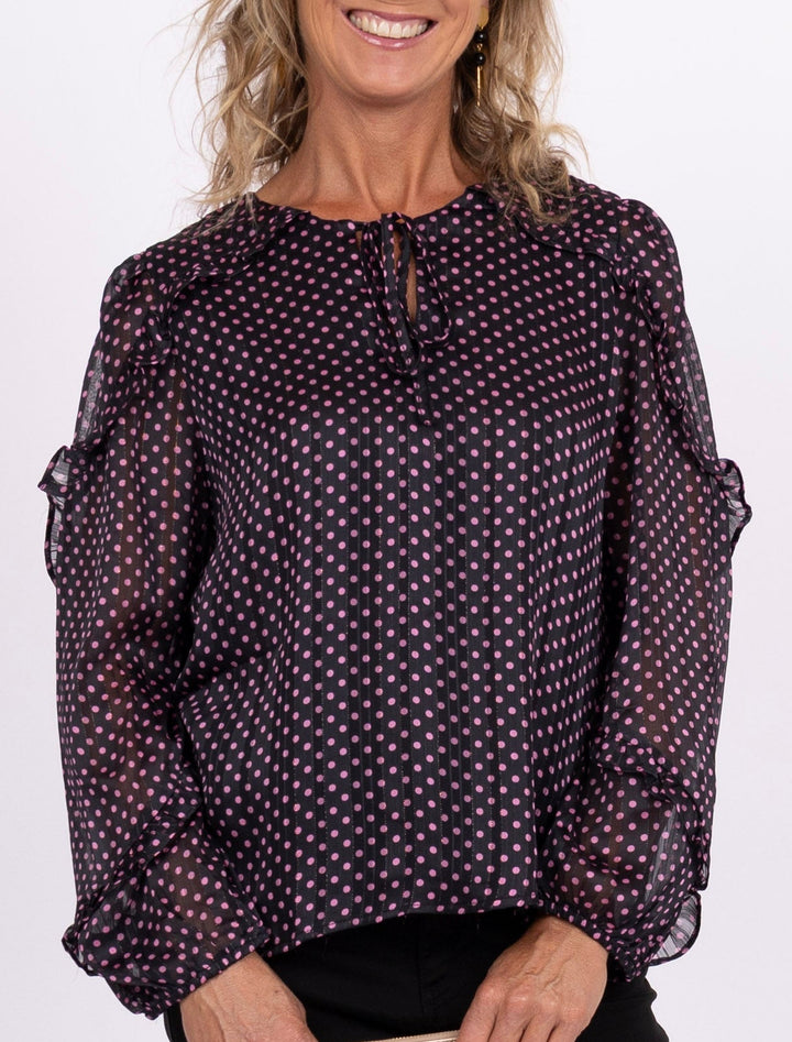 A smiling woman stands against a plain light background wearing a sheer long-sleeve blouse with a small repeating pattern, black pants, and pink sneakers, while holding a small cream clutch.