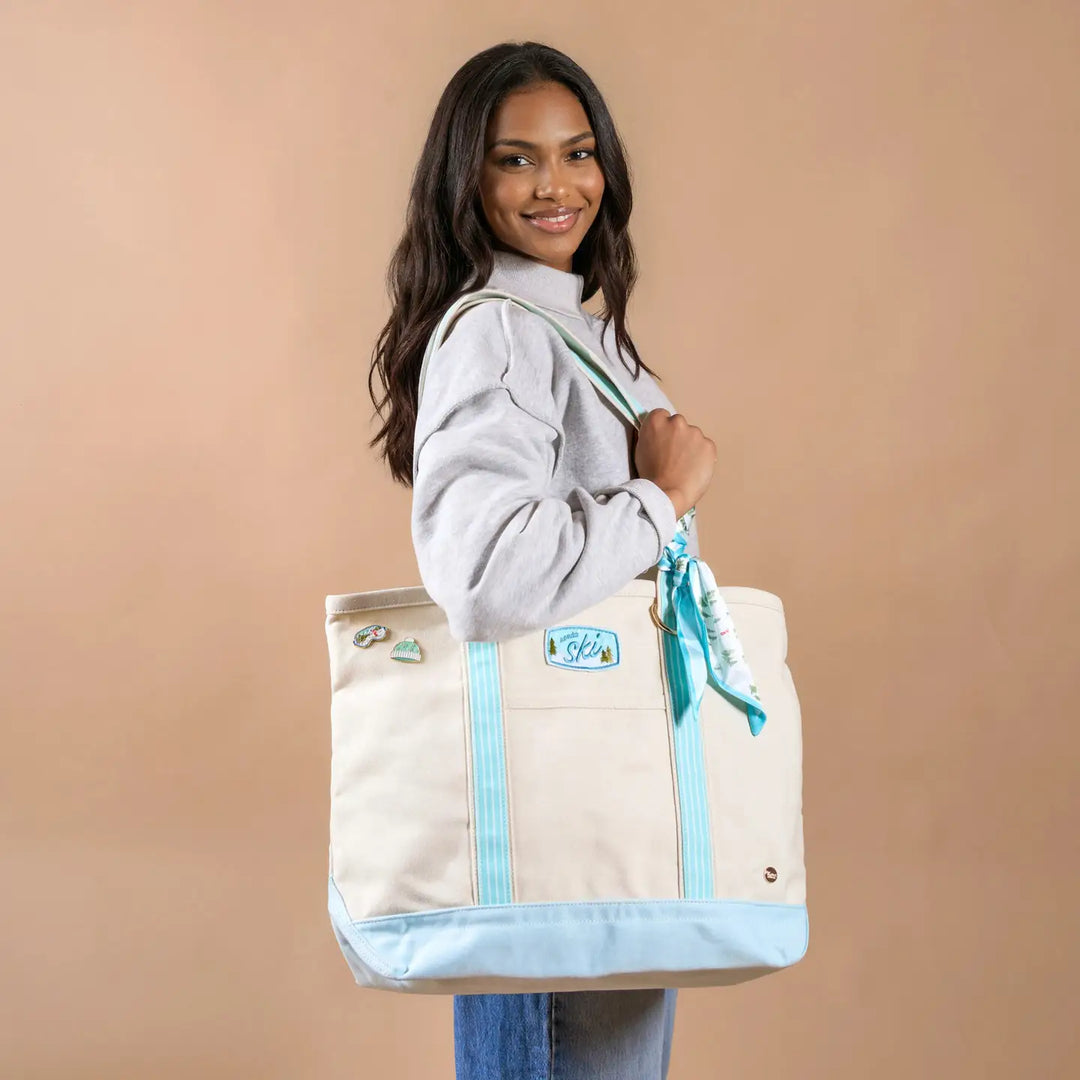 A woman wearing a gray sweatshirt and blue jeans carries a large cream canvas tote bag with light blue trim and a matching light blue scarf tied to the handle, set against a solid beige background.