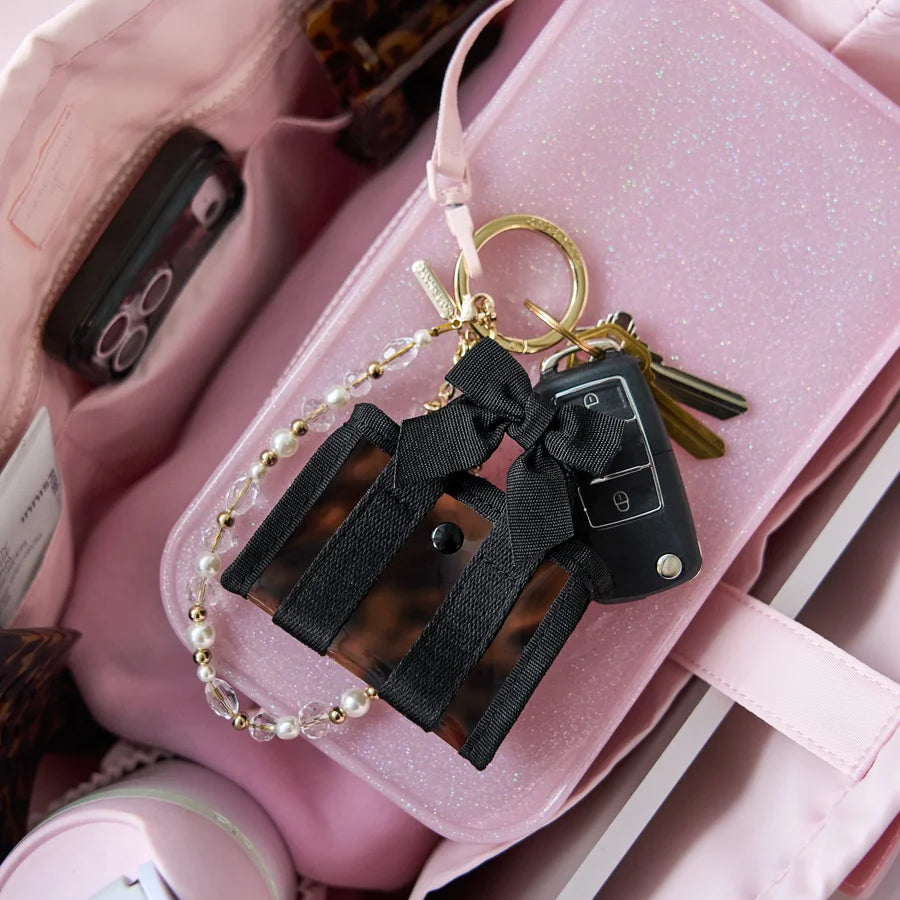 A pink and black keychain bag charm with a leopard print body, black ribbon bow, gold keyring, and pearl chain, shown inside a pink bag.