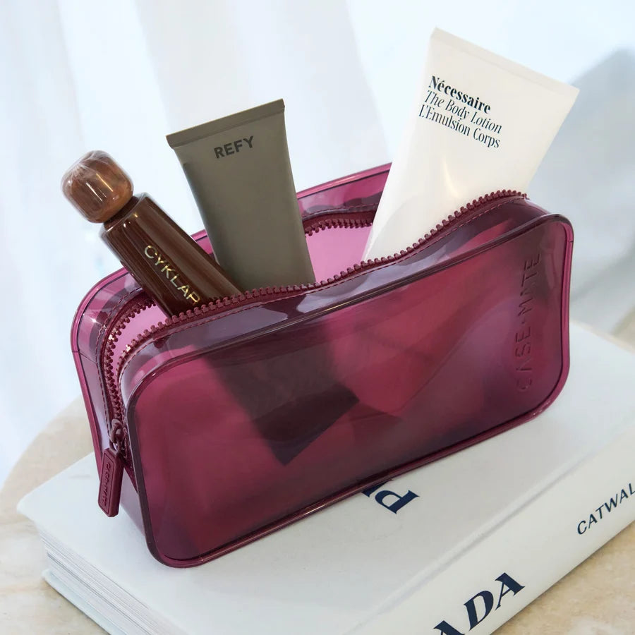 A translucent magenta cosmetic pouch filled with skincare items, including a white tube labeled Nécessaire and a brown bottle labeled CYKLAR, styled on top of a white book.