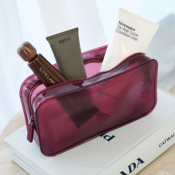 A translucent magenta cosmetic pouch filled with skincare items, including a white tube labeled Nécessaire and a brown bottle labeled CYKLAR, styled on top of a white book.