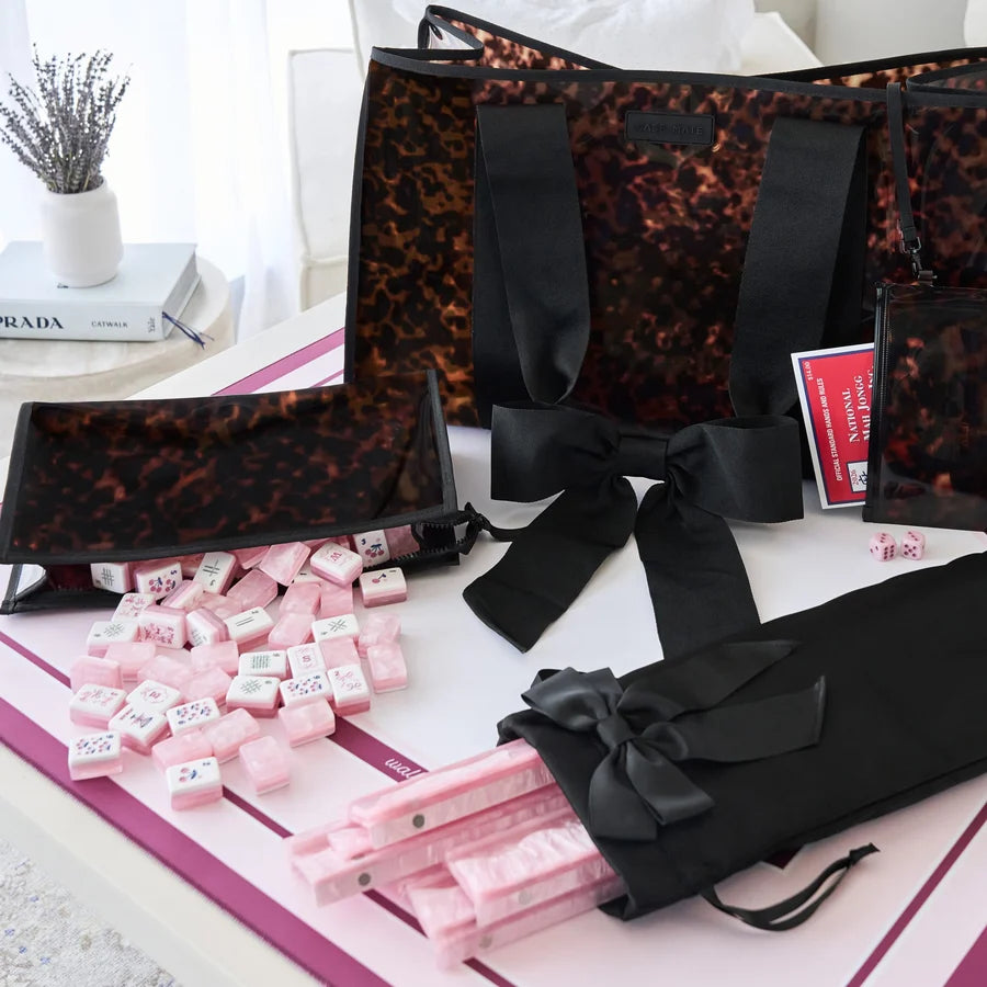 A coordinated gift set featuring a large tote bag, smaller matching pouches, a black ribbon bow, pink wrapped items, and pink dice, arranged on a white surface with a pink-and-white mat. A small vase with dried stems and a book are visible in the background.