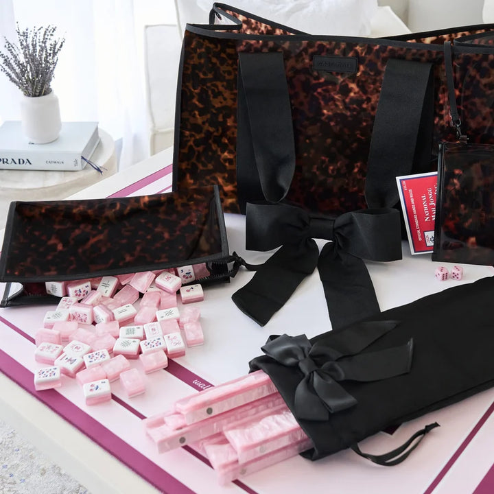 A coordinated gift set featuring a large tote bag, smaller matching pouches, a black ribbon bow, pink wrapped items, and pink dice, arranged on a white surface with a pink-and-white mat. A small vase with dried stems and a book are visible in the background.