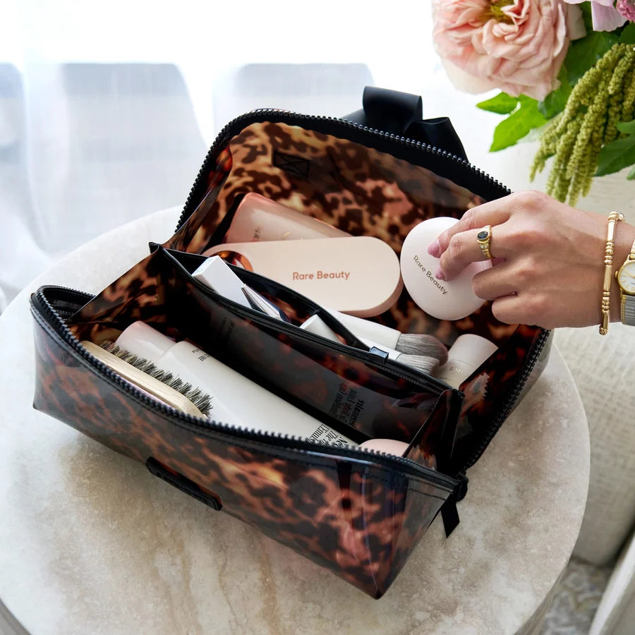 A leopard-print cosmetic bag filled with beauty products, shown open on a round table with a hand reaching in. The bag has a translucent leopard pattern with black trim, and the scene includes soft-focus flowers in the background.