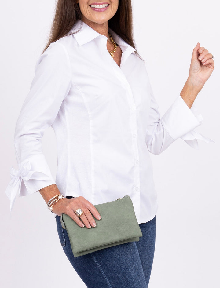 A woman modeling a white long-sleeve button-up shirt, dark blue flared jeans, and brown clogs, holding an olive green clutch against a plain white studio background.