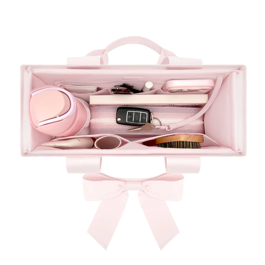 A light pink vanity organizer case with a top handle and bow, shown open and filled with beauty and travel accessories on a white background.