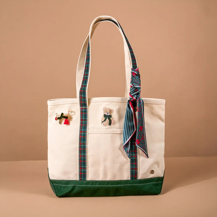A cream canvas tote bag with a dark green bottom panel, featuring two shoulder straps with a red and green plaid pattern, a matching patterned scarf tied to the side, and embroidered teddy bear and bow details on the front pockets.