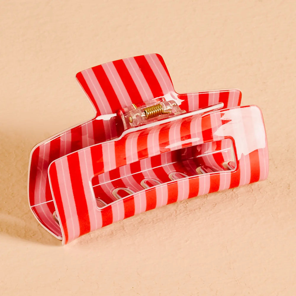 A glossy hair claw clip with alternating red and light pink stripes, shown on a textured beige background.