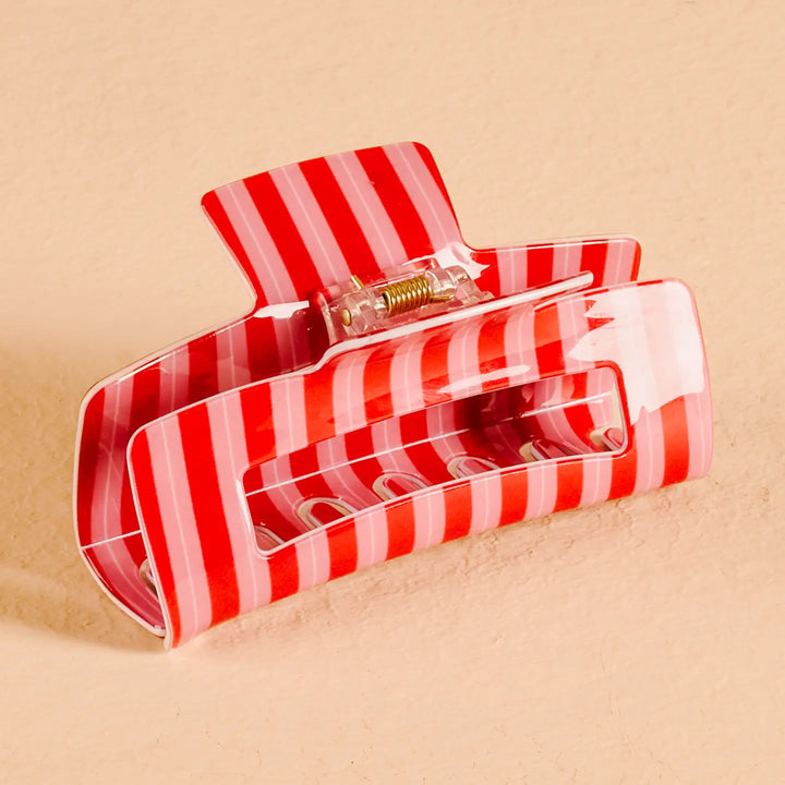 A glossy hair claw clip with alternating red and light pink stripes, shown on a textured beige background.
