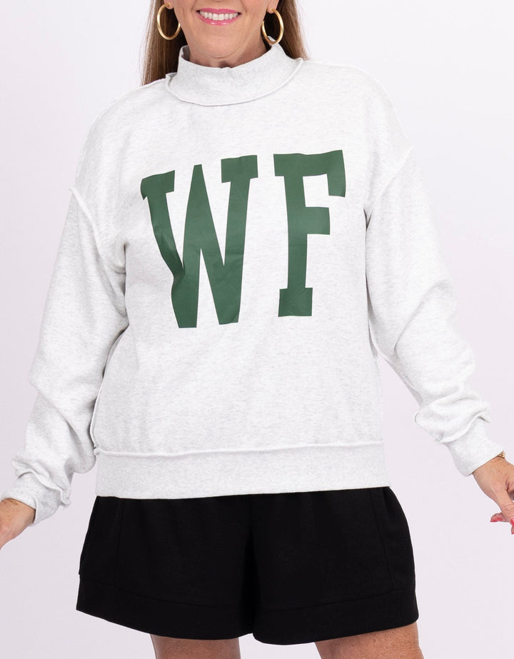 A woman wearing a white mock-neck sweatshirt with large green block letters &