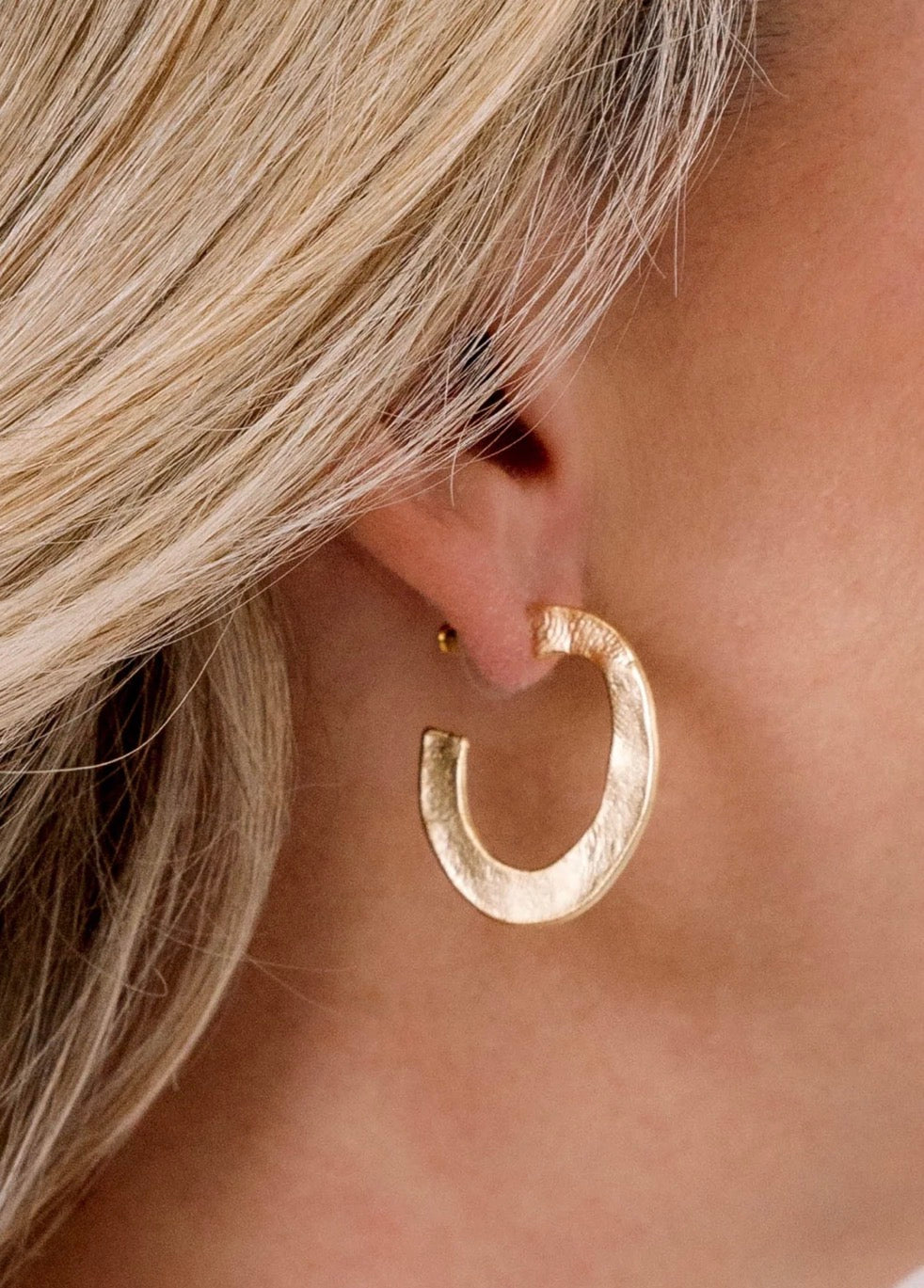 A close-up of a model's ear wearing a chunky, textured gold crescent hoop earring.
