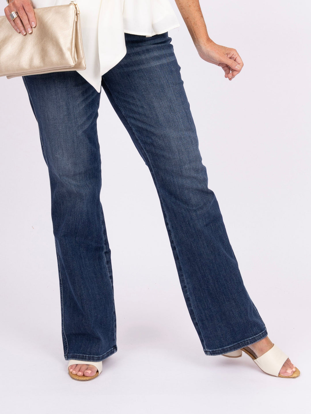 A woman modeling a sleeveless cream wrap-style top with a tie waist, paired with dark blue flare jeans, a metallic gold clutch, and white heeled sandals, against a plain white studio background.