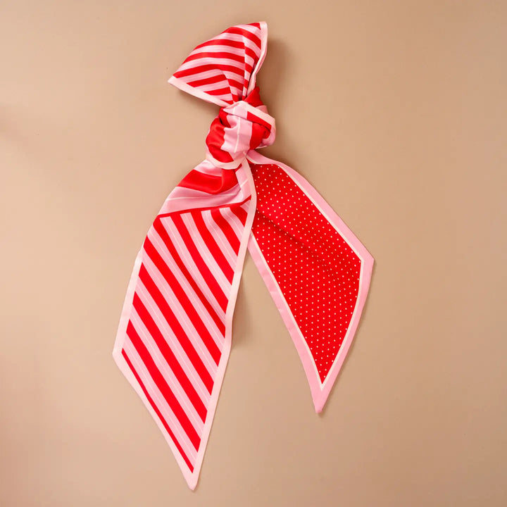 A red and white patterned scarf tied in a knot, displayed on a solid beige background. One side of the scarf features red and white diagonal stripes, while the other side features a red background with small white polka dots and a light pink border.